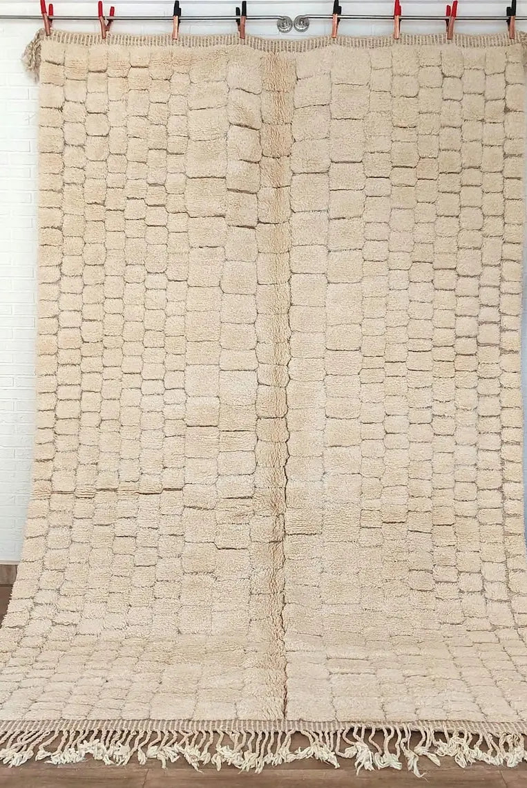 Moroccan beni mrirt rug Beige woven with tassels on a wooden floor with a vase and plant in the background.