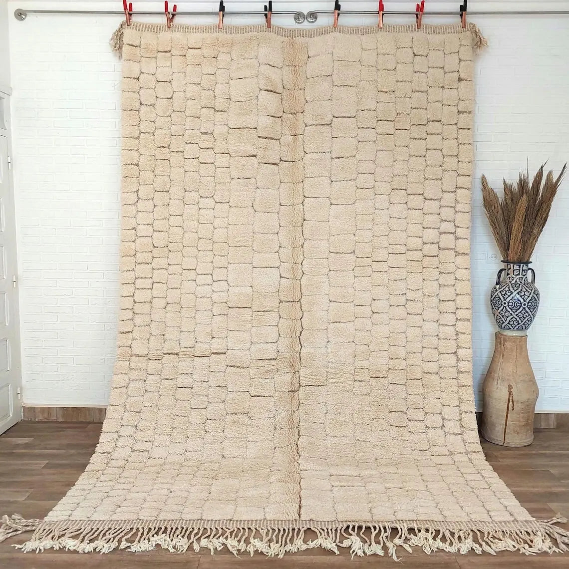 Moroccan beni mrirt rug Beige woven with tassels on a wooden floor with a vase and plant in the background.