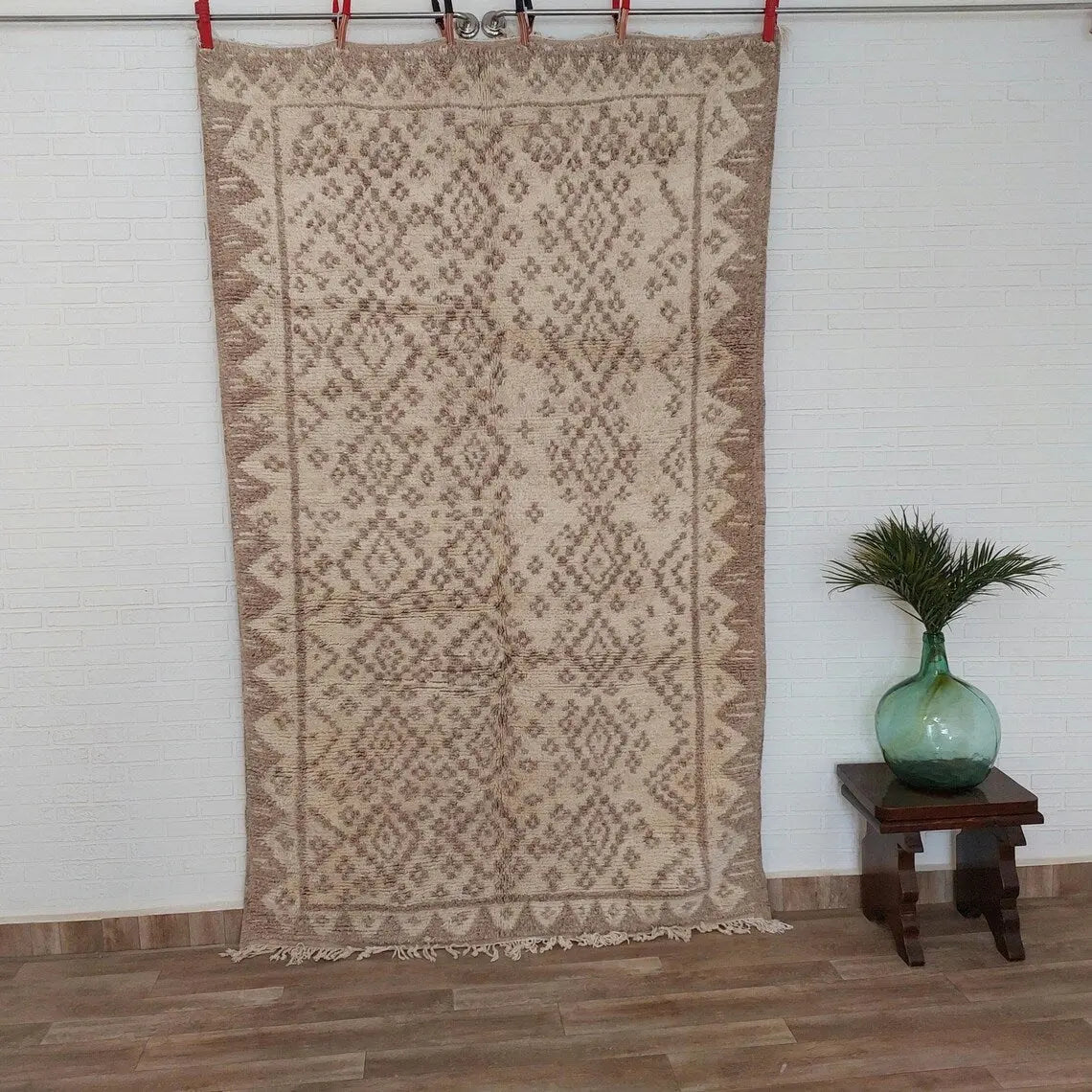 Taznakht Beige patterned rug hanging on a rack with a small table and green vase in the background.
