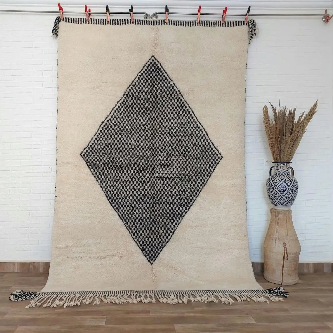 Beige beni mrirt rug with black diamond pattern hanging on a rod, with a vase and dried plants in the background.