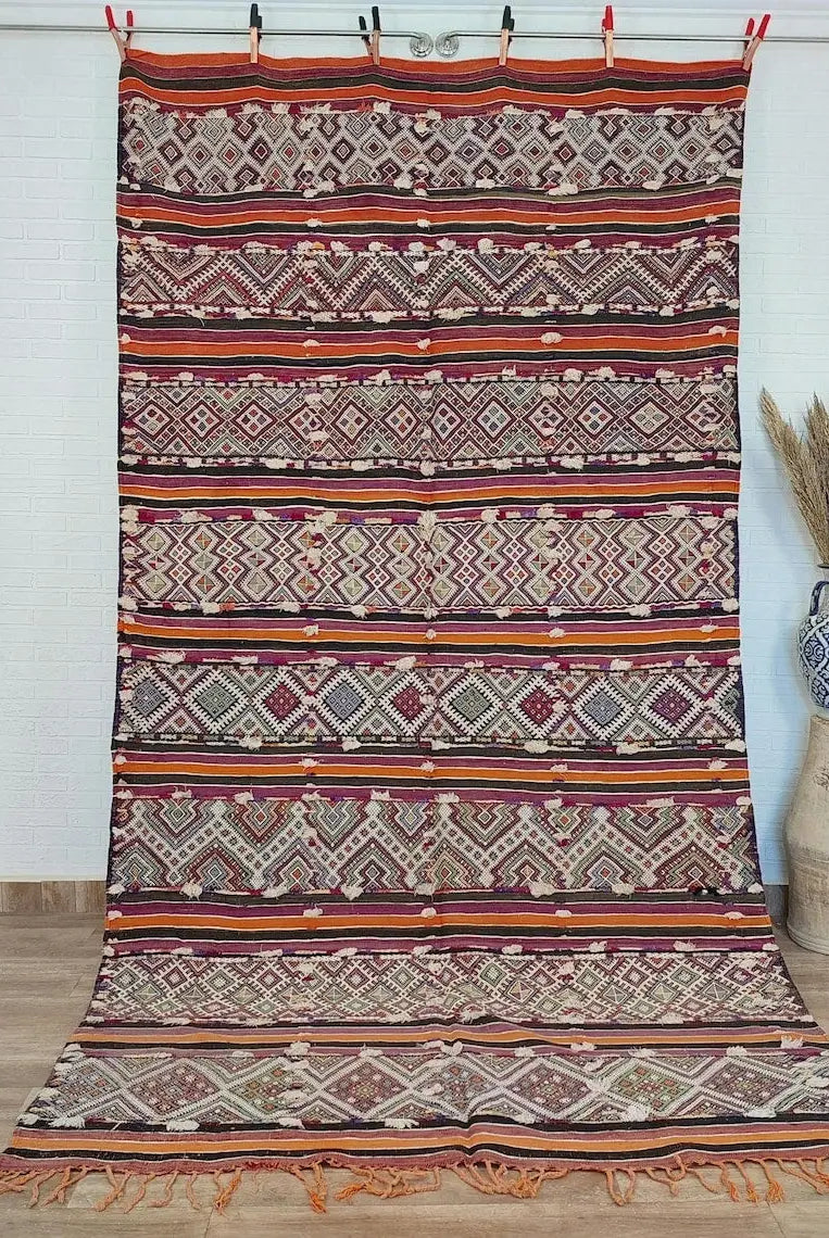 Colorful patterned Moroccan rug on a wooden floor with a vase and dried plants in the background.