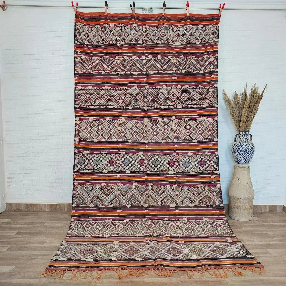 Colorful patterned Moroccan rug on a wooden floor with a vase and dried plants in the background.